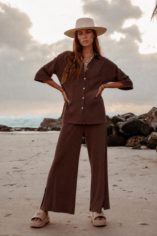 Woman in a brown outfit with a hat standing on a beach with rocks and ocean in the background