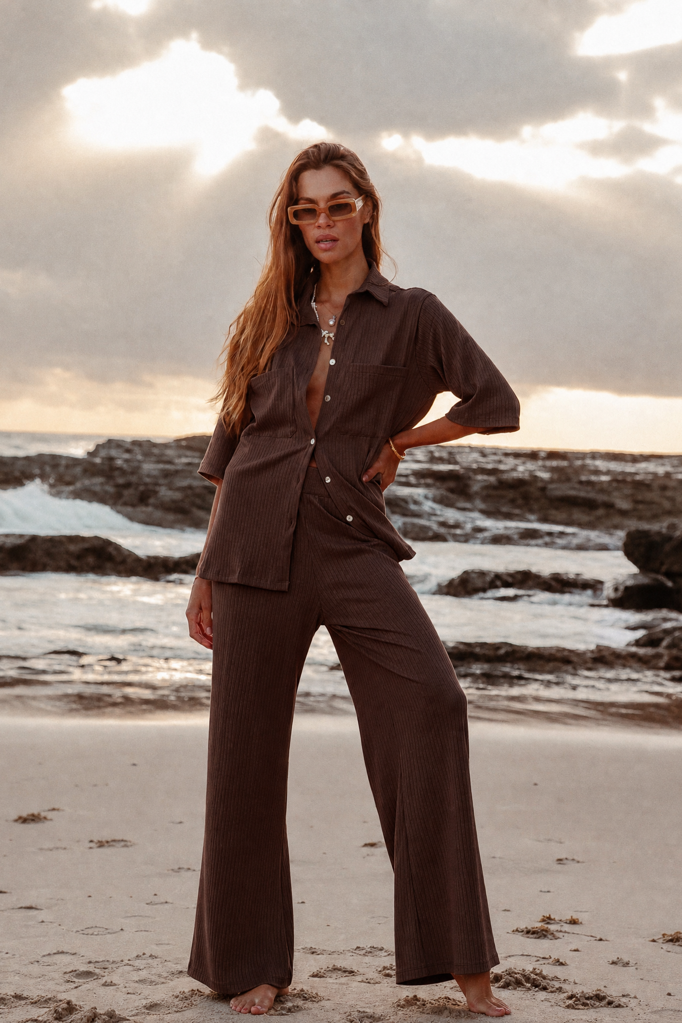 Woman in a brown outfit standing on a beach with a sunset in the background